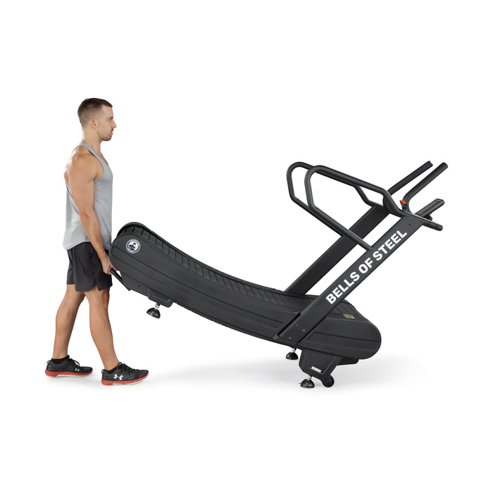 A man in athletic wear tilts and rolls the Blitz Magnetic Resistance Manual Treadmill by Bells of Steel using its rear wheels. The black, curved treadmill features handlebars and is angled upwards as he moves this sled push treadmill.