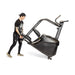 A person tilts and moves the Bells of Steel Blitz Stepper—a large curved treadmill with handles and wheels—on a white background, designed for low-impact cardio workouts.