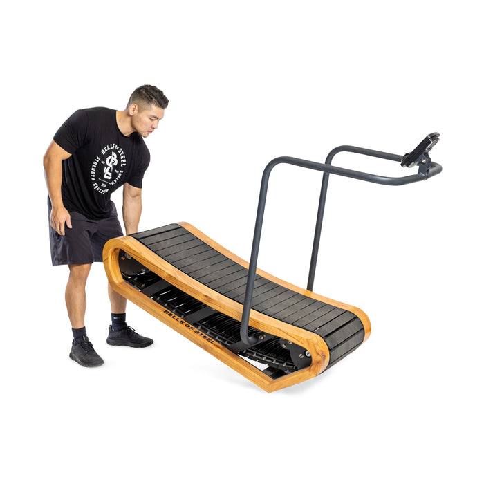 A person in a black t-shirt and shorts adjusts the Bells of Steel Wooden Residential Manual Treadmill, featuring a sleek black surface, wooden frame, and minimalist design with a handlebar device holder.