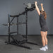 A woman in athletic wear stands before the Bells of Steel USA Pandemonium Squat – 3-in-1 Pendulum Squat / Calf Press / Viking Press Machine, reaching up to grip the handles as she prepares for an overhead exercise.