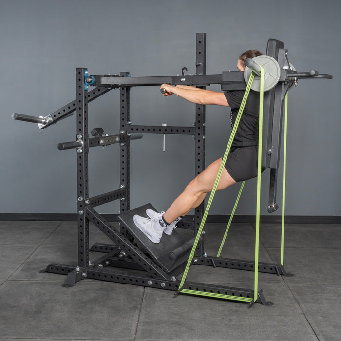 A person prepares to squat in a gym using the Bells of Steel USA Pandemonium Squat – a 3-in-1 pendulum squat, calf press, and Viking press machine – standing on its slanted platform and gripping the handles.