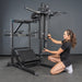 A woman in athletic wear kneels beside the Bells of Steel USA Pandemonium Squat – 3-in-1 Pendulum Squat / Calf Press / Viking Press Machine, adjusting a padded bar. The equipment is set against a gray wall on a concrete gym floor.